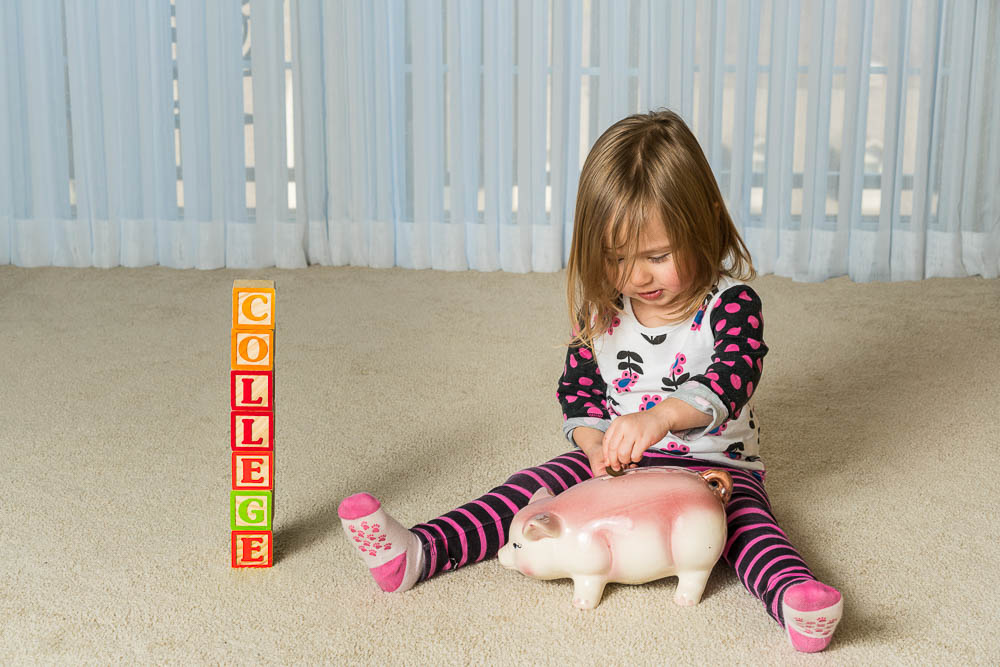 young girl on the floor with a piggy bank and blocks arranged to spell college